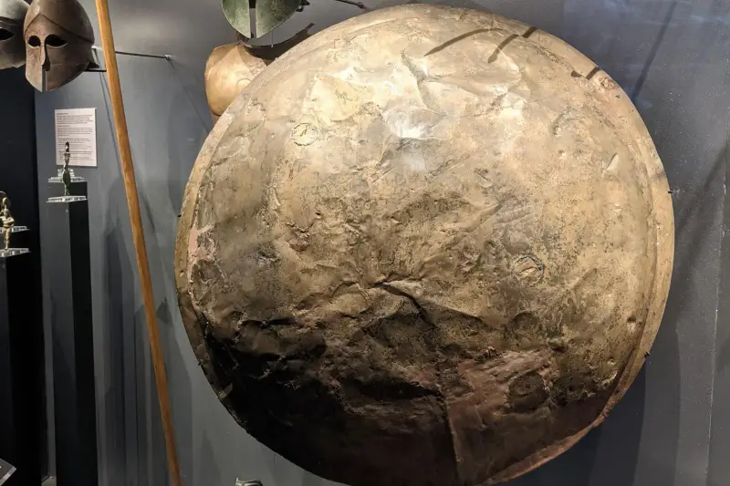 A museum display case of ancient bronze helmets, a large round shield, and spears at the Thermopylae Museum