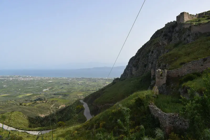 The fortress walls and a tower of Acrocorinth on the rocky ridge, with the coastal plain and the Gulf of Corinth spread out below
