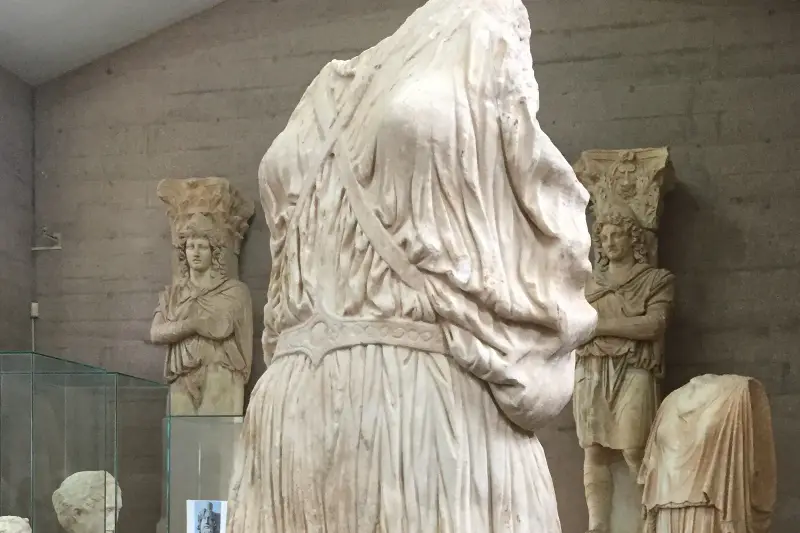 A large draped marble torso filling the frame, cropped above the shoulders, flanked by two carved figures carrying basket capitals on their heads and a headless torso at the right, against the museum's concrete wall